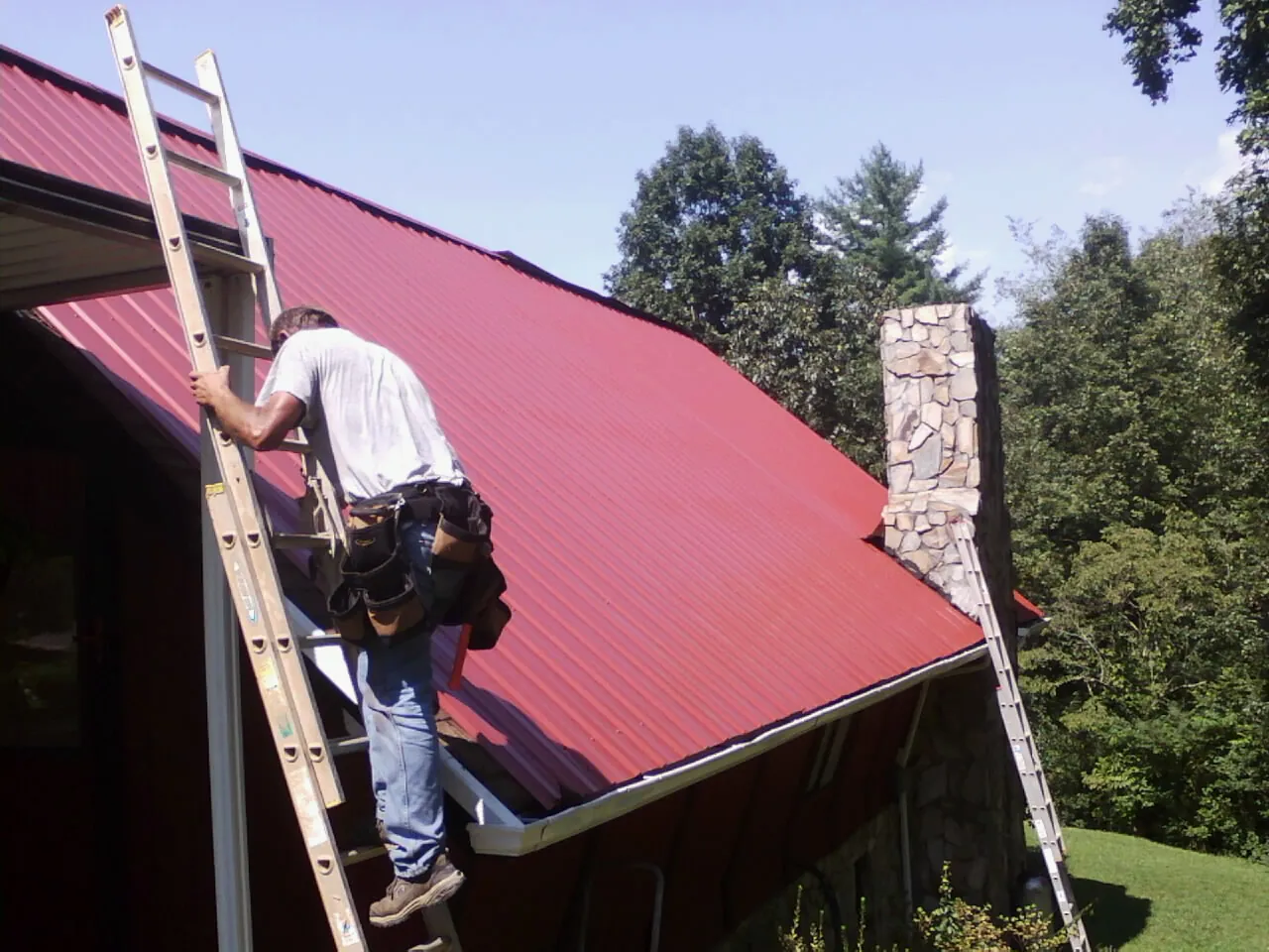 Roofer on a ladder beside a red metal roof and stone chimney — Soffit & Fascia Repair by Roofing Brothers in Bethel