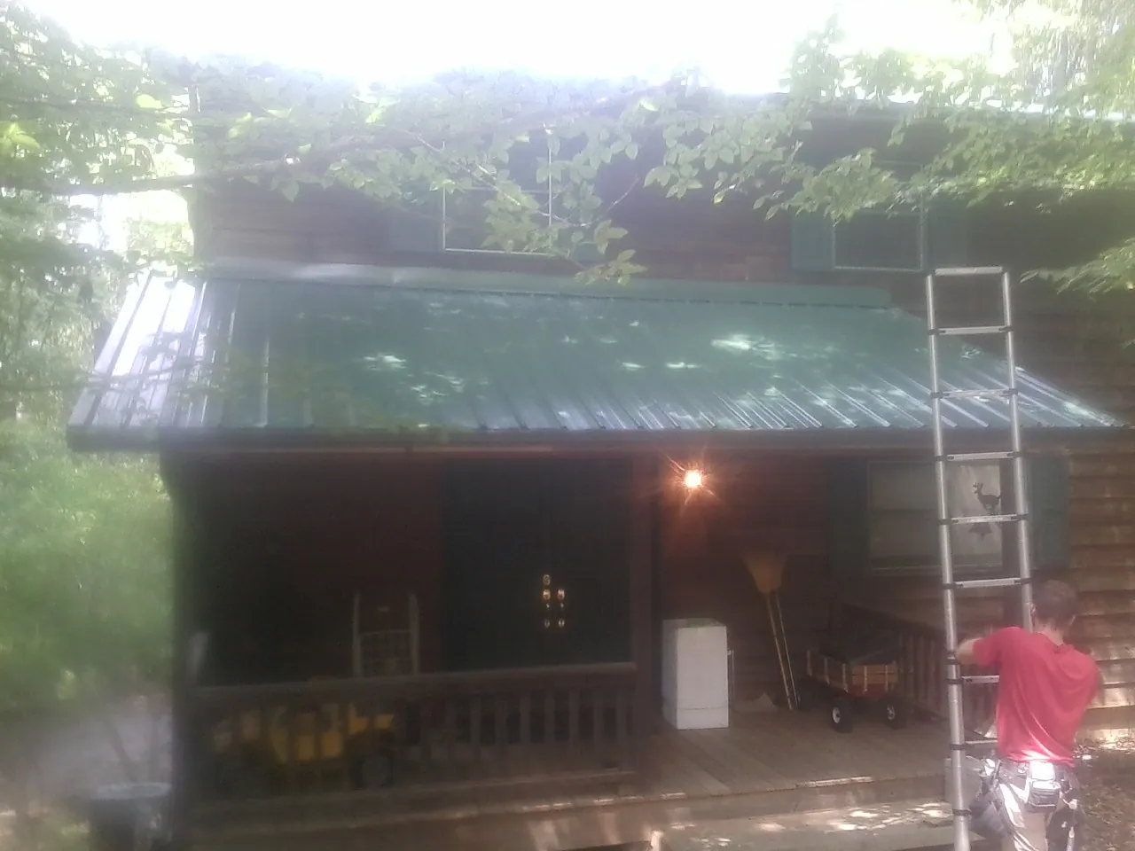 Worker on a ladder at the green porch roof of a wooded cabin in Bethel